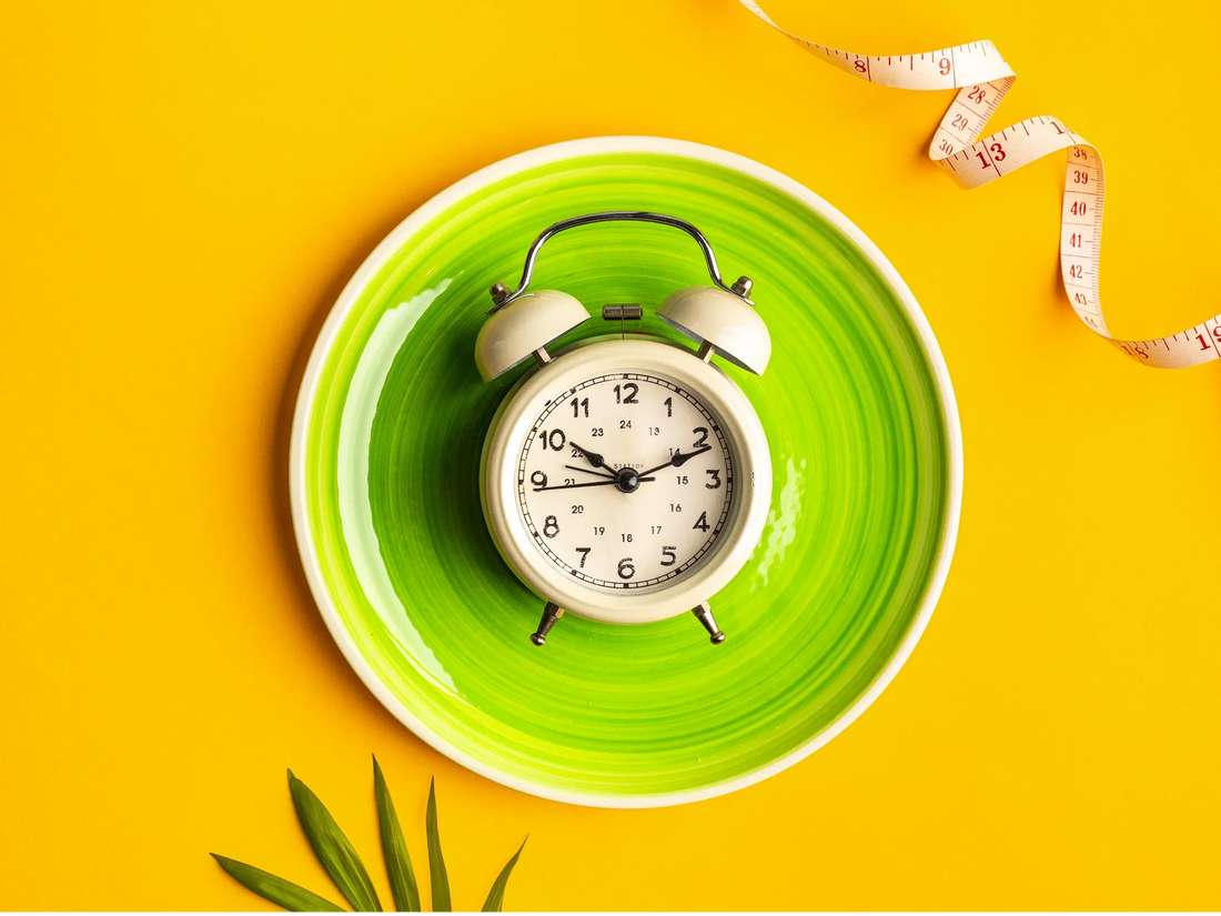 An alarm clock placed on a green plate next to a measuring tape on a yellow background, symbolizing time-restricted eating and the concept of intermittent fasting schedules for weight management